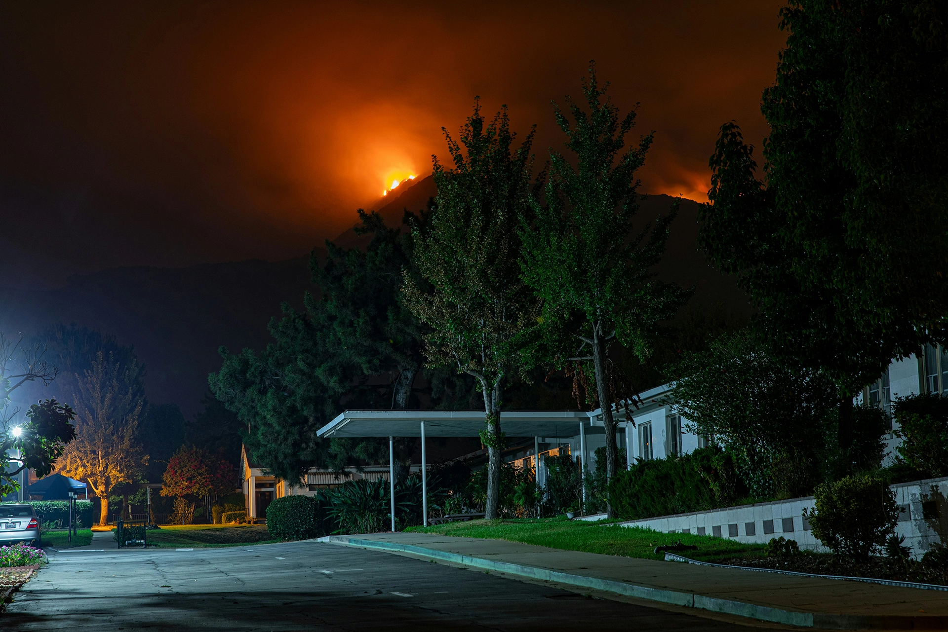 A residential street at night with a wildfire blazing on a nearby hillside. The fire illuminates the sky with a bright orange glow, creating a stark contrast against the dark surroundings.