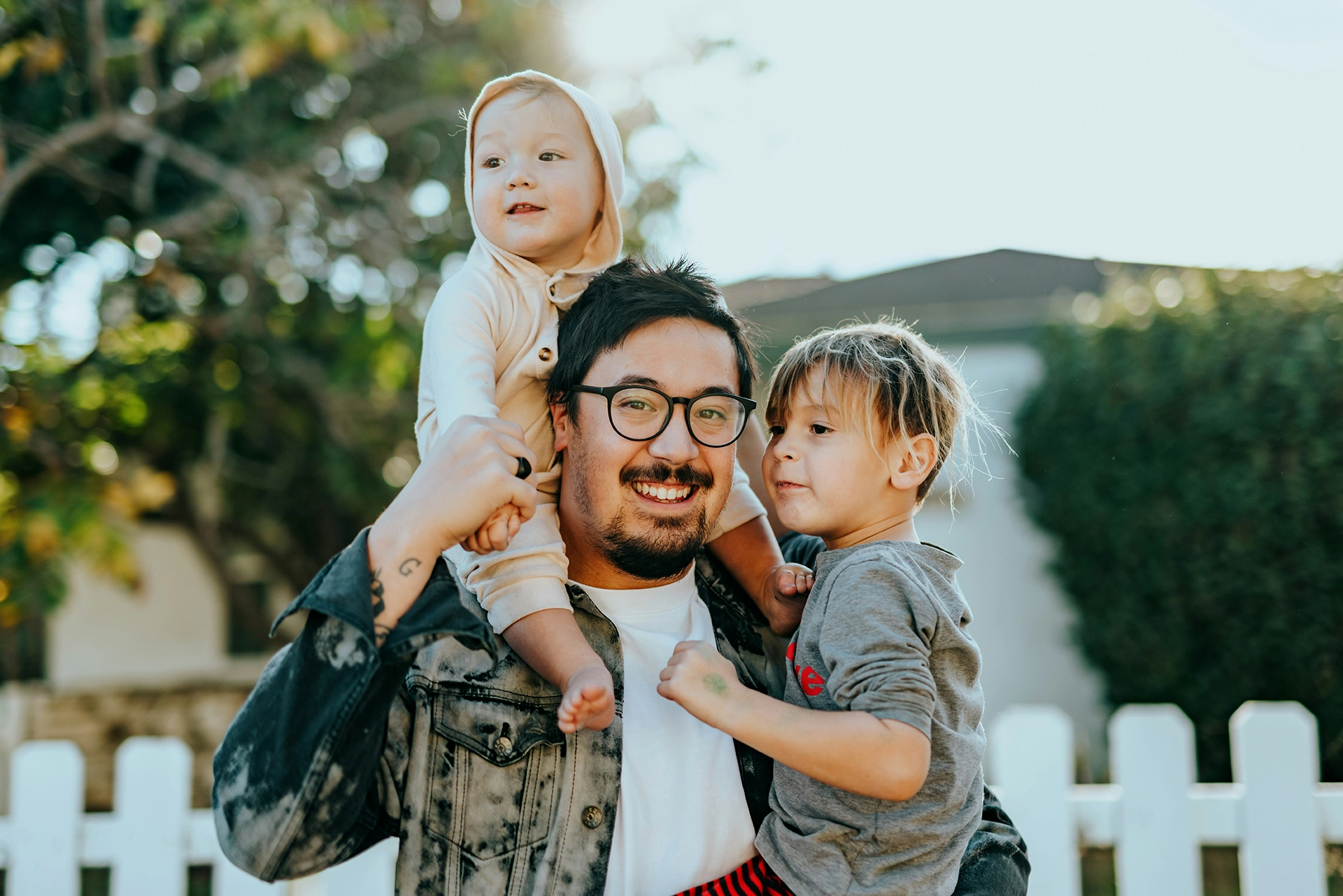 A father wearing glasses smiling and holding two young children, one sitting on his shoulders and the other in his arms, outdoors near a white picket fence.