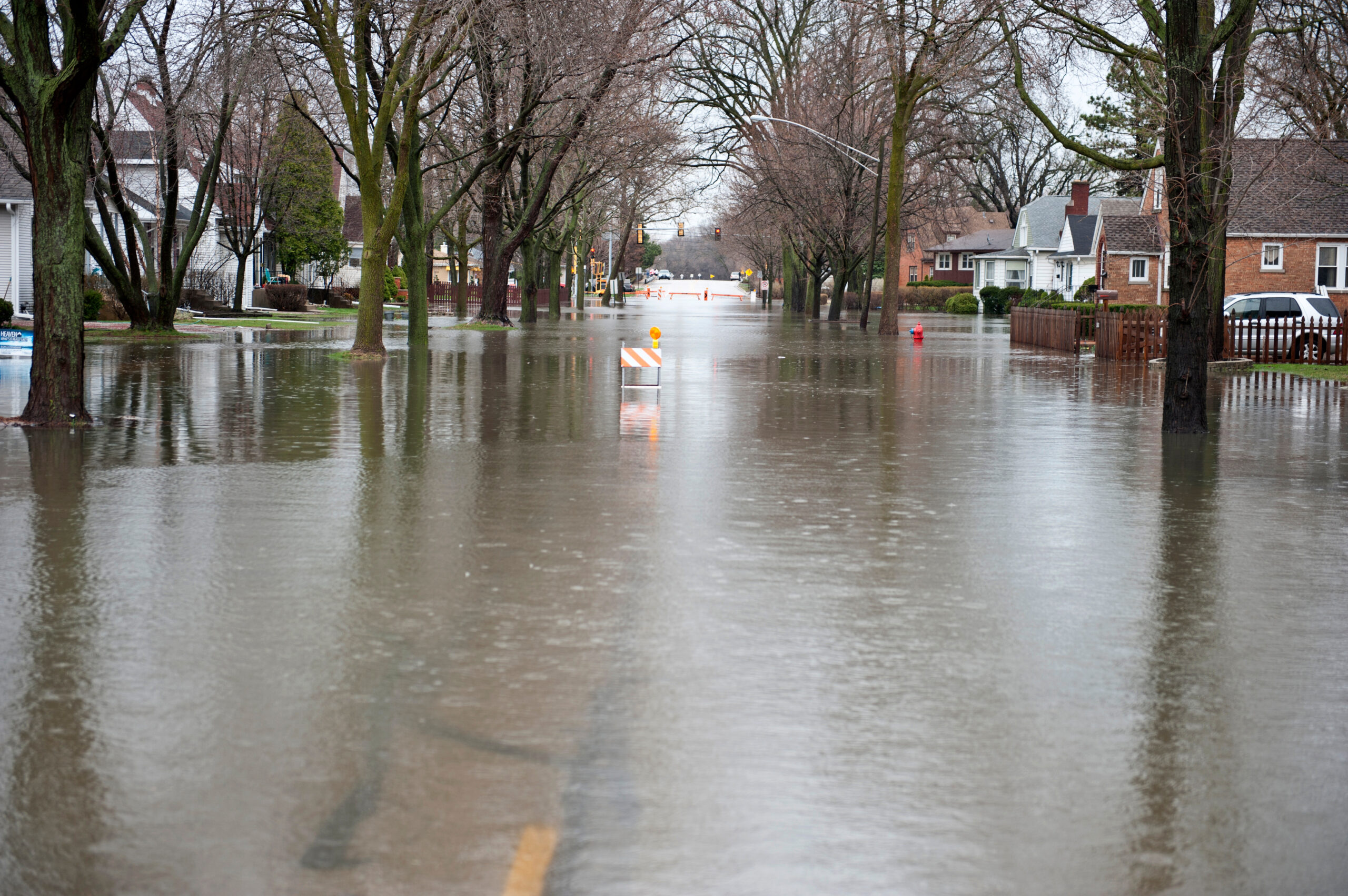 A flooded residential street lined with trees and houses, with deep water covering the road and traffic barricades partially submerged in the distance.