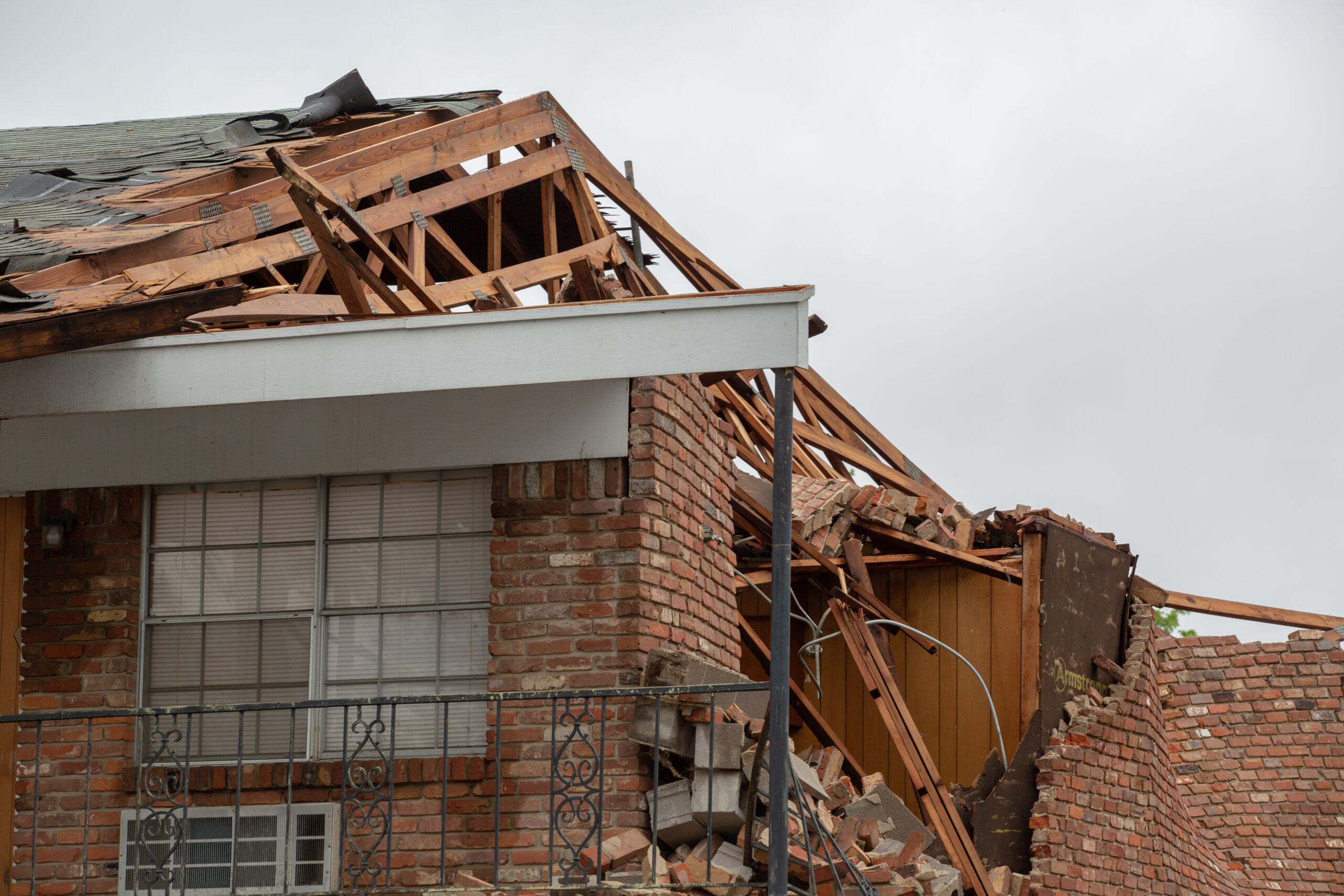 A brick house with extensive roof damage, showing exposed wooden framing and debris from a collapsed structure.