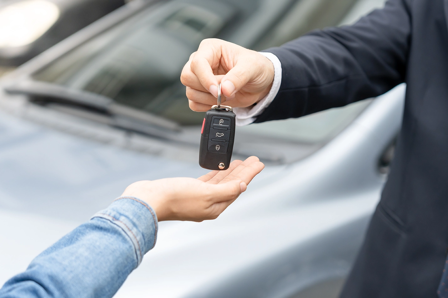 A close-up of a car key being handed over from one person to another in front of a parked car.